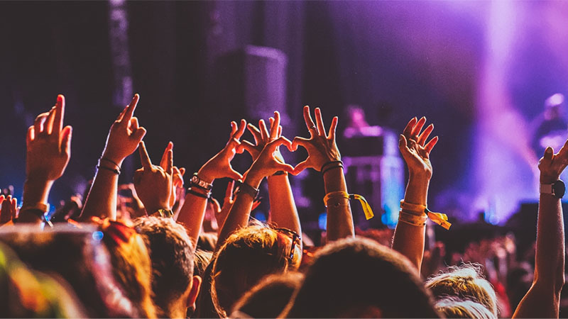 A crowd at a concert raising their hands, some forming heart shapes, against a backdrop of colorful stage lighting.